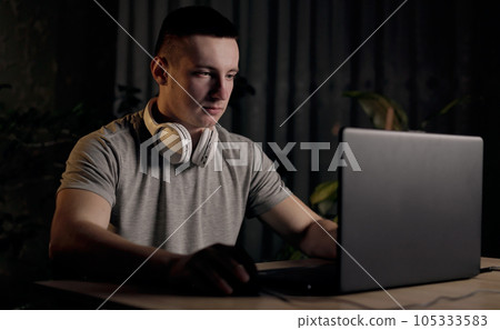 Young man freelancer using laptop, typing, scrolling, surfing web, looking at screen at night at home. Male smiling and communication on social media, video conference call. Distance learning online 105333583
