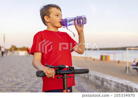 A boy drinks water from plastic bottle, standing with scooter 105334970