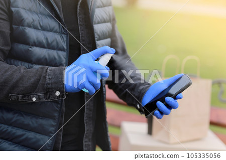 Delivery service courier during the Coronavirus, COVID-19, pandemic, cropped courier hands in gloves spraying alcohol disinfectant spray on cellphone near cardboard boxes outdoors. Delivery service courier during the Coronavirus, COVID-19, pandemic, cropped courier hands in gloves spraying alcohol disinfectant spray on cellphone near cardboard boxes outdoors. 105335056