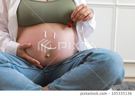 Close-up of torso of young pregnant model applying moisturizer on her belly to avoid stretch marks. Future mom with funny smile from moisturizing cream on her tummy. Pregnancy skincare concept 105335136