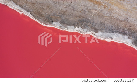 Flying over a pink salt lake. Salt production facilities saline evaporation pond fields in the salty lake. Dunaliella salina impart a red, pink water in mineral lake with dry cristallized salty coast. 105335137