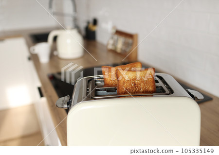 Toasted bread and toaster for breakfast. Modern white toaster and roasted bread slices toasts inside on wooden table in kitchen. 105335149