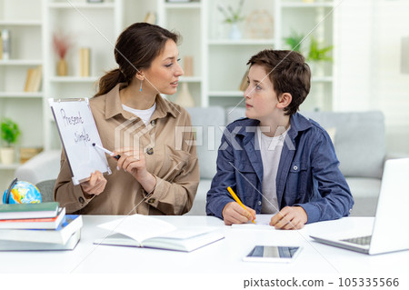 Home education, woman teaches boy at home sitting at table in living room, mother explains educational material to son doing homework together. Home education, woman teaches boy at home sitting at table in living room, mother explains educational material to son doing homework together. 105335566