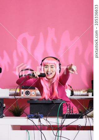 Woman performer sitting at dj table performing techno music using professional mixer console, wearing headset and talking with fans using microphone. Asian artist playing electronic song at night 105336633