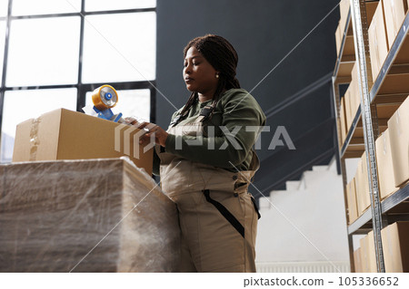 Worker using adhesive tape to pack clients orders, preparing products for shipping in storage room. African american employee wearing industrial overall working at goods inventory in warehouse 105336652