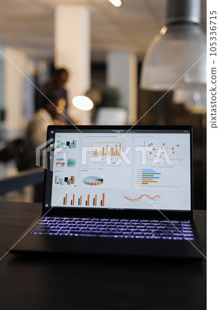Laptop computer with marketing statistics graphs on screen, standing on table in startup office, coworkers discussing company strategy in background. Employees working overtime in startup office 105336715