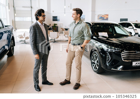 Full length portrait of smiling male client talking to car dealer wearing business suit in dealership discussing automobiles looking at luxurious new model. Concept of choosing and buying new auto Full length portrait of smiling male client talking to car dealer wearing business suit in dealership discussing automobiles looking at luxurious new model. Concept of choosing and buying new auto 105346325