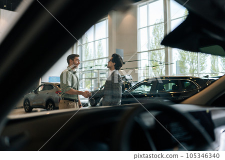 Remote view from inside of car through windshield to handsome smiling car dealer talking with cheerful male in showroom and shaking hand. Cheerful two people standing in dealership and talking. 105346340