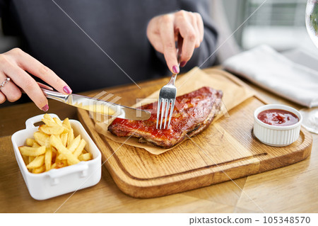 Lunch in a restaurant, a woman cuts Delicious Pork ribs. Full rack of ribs BBQ on wooden plate with french fries and salad Lunch in a restaurant, a woman cuts Delicious Pork ribs. Full rack of ribs BBQ on wooden plate with french fries and salad 105348570