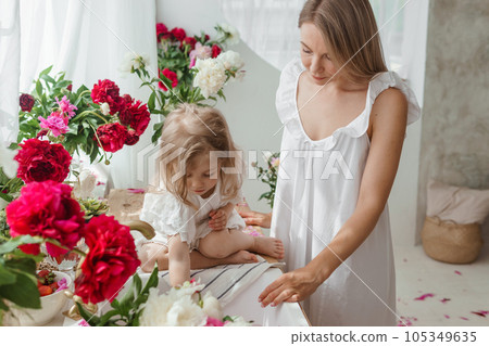 A little blonde girl with her mom on a kitchen countertop decorated with peonies. The concept of the relationship between mother and daughter. Spring atmosphere. A little blonde girl with her mom on a kitchen countertop decorated with peonies. The concept of the relationship between mother and daughter. Spring atmosphere. 105349635