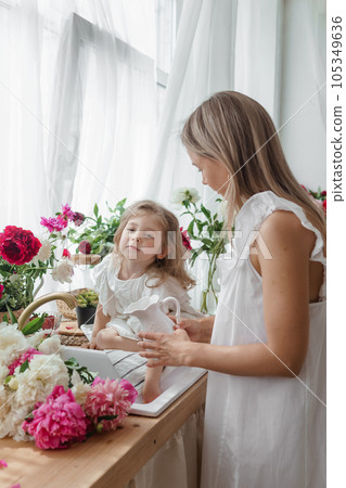 A little blonde girl with her mom on a kitchen countertop decorated with peonies. The concept of the relationship between mother and daughter. Spring atmosphere. A little blonde girl with her mom on a kitchen countertop decorated with peonies. The concept of the relationship between mother and daughter. Spring atmosphere. 105349636