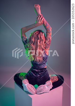 Young fitness blonde woman wearing blue velour booty shorts posing on camera, sits on top of big cube, back view Young fitness blonde woman wearing blue velour booty shorts posing on camera, sits on top of big cube, back view 105352623