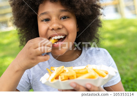 Dark skinned female child eating junk food with pleasure in park while sitting on green grass. 105353114