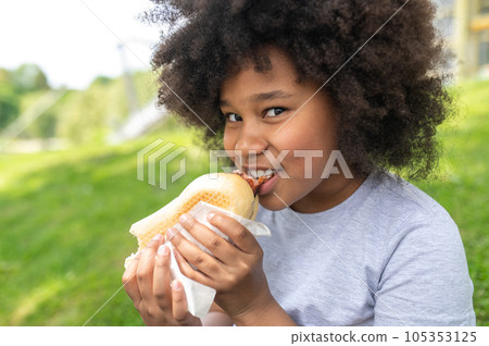Dark skinned female child eating junk food with pleasure in park while sitting on green grass. 105353125