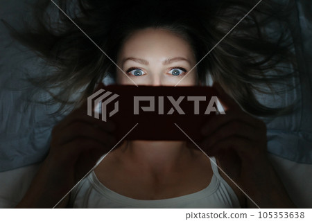 Young female addictively staring at her smartphone late at night in bed. 105353638