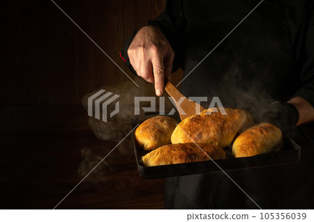 The baker bakes hot dog buns. Kitchen scraper in the chef hand. Space for advertising on a dark background 105356039