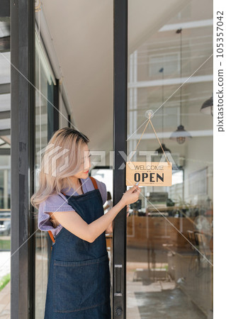 Young asian businesswoman small business owner standing at door entrance signboard, A cheerful entrepreneur young waitress in a blue apron near the glass door Young asian businesswoman small business owner standing at door entrance signboard, A cheerful entrepreneur young waitress in a blue apron near the glass door 105357042