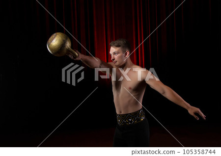 Power gymnast circus with weight.Handsome guy with a muscular body holding a kettlebell on a black stage background 105358744