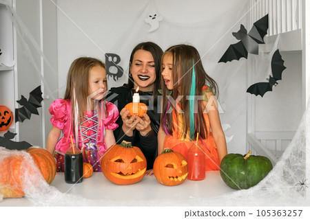 Dressed up in carnival costumes, Daughters and mom blow out a candle on a pumpkin on the eve of Halloween. The room is decorated with paper bats, cobwebs and carved Jack-o'-lantern pumpkins Dressed up in carnival costumes, Daughters and mom blow out a candle on a pumpkin on the eve of Halloween. The room is decorated with paper bats, cobwebs and carved Jack-o'-lantern pumpkins 105363257