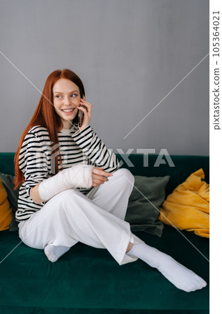 Vertical portrait of positive young woman with broken arm wrapped in gypsum bandage talking on smartphone with friends during rehabilitation. Smiling female with injured hand from accident at home. 105364021