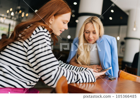 Side view of two attractive girlfriends women sitting in cafe looking at old photos on smartphone and smiling. Best females friends sitting at coffee shop looking at phone. Concept of woman friendship Side view of two attractive girlfriends women sitting in cafe looking at old photos on smartphone and smiling. Best females friends sitting at coffee shop looking at phone. Concept of woman friendship 105366485