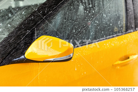 Front side mirror of bright yellow car washed in self serve carwash, water spraying over glass 105368257