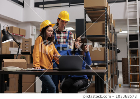 Asian distribution center workers checking goods supply schedule on laptop. Warehouse woman manager sitting at desk while explaining logistics chain to coworkers in industrial storage room 105368578