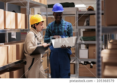 Man and woman warehouse coworkers supervising inventory together and reading report on laptop. Diverse employees working in stockroom and recording storehouse activity on computer Man and woman warehouse coworkers supervising inventory together and reading report on laptop. Diverse employees working in stockroom and recording storehouse activity on computer 105368579
