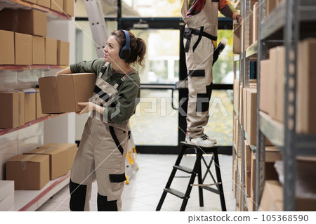 Supervisor looking at cardboard boxes in storage room, listening music at headset while preparing clients orders in storehouse. Executive manager working at products delivery in warehouse Supervisor looking at cardboard boxes in storage room, listening music at headset while preparing clients orders in storehouse. Executive manager working at products delivery in warehouse 105368590