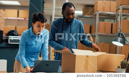 Team of entrepreneurs packing order for shipment, using cardboard boxes and warehouse stock. Cheerful people working on supply chain and distribution logistics, quality control. Handheld shot. 105368598