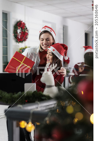 Exuberant worker receiving gift from colleague during Christmas holiday season. Excited employee gets red box present from coworker on secret santa party in festive decorated workspace 105368646
