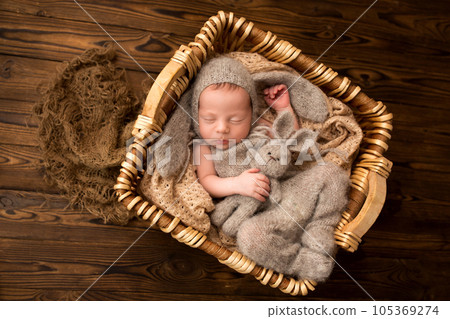 A cute newborn baby boy sleeps in a gray overalls in the first days of life. Against a background of dark brown wood. In a wicker wooden basket. Bunny gray felt toy. Professional photography, portrait 105369274