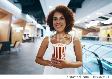Curly-haired woman with an afro hairstyle in a white shirt is walking to watch a movie with popcorn in hand Curly-haired woman with an afro hairstyle in a white shirt is walking to watch a movie with popcorn in hand 105369321