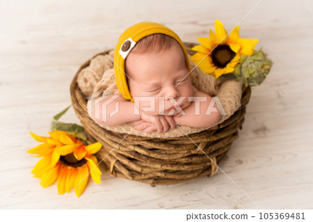 A cute newborn boy sleeps in a yellow-orange hat in the first days of life. Against a background of light white wood. In wicker wooden basket. With sunflower flowers. Professional photography portrait A cute newborn boy sleeps in a yellow-orange hat in the first days of life. Against a background of light white wood. In wicker wooden basket. With sunflower flowers. Professional photography portrait 105369481