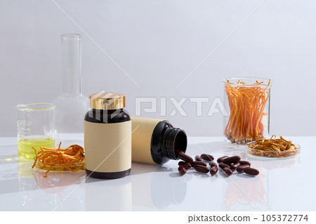 Two medicine bottle unbranded with capsules decorated with glassware containing cordyceps and yellow liquid on white background. Scene for advertising product. Space for design Two medicine bottle unbranded with capsules decorated with glassware containing cordyceps and yellow liquid on white background. Scene for advertising product. Space for design 105372774