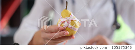 Nutritionist holds pear fruit and measuring tape in his hand and advises to eat healthy food with vitamins. Nutritionist holds pear fruit and measuring tape in his hand and advises to eat healthy food with vitamins. 105375346