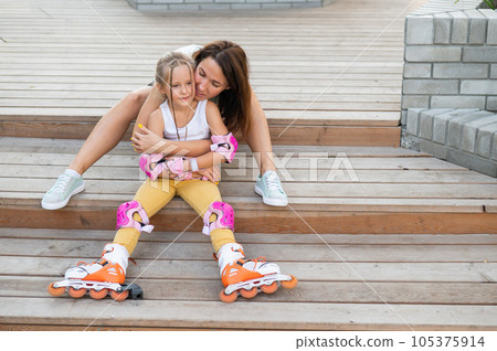 Little girl in roller skates and her mom sit on a wooden ladder and hug outdoors.  105375914
