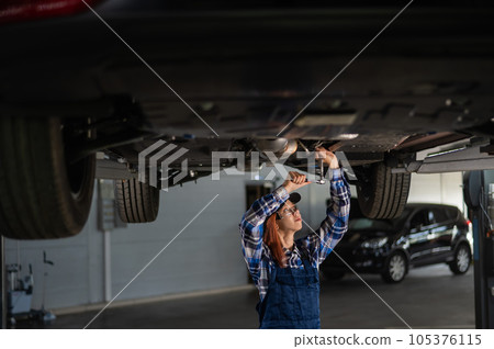 Female mechanic unscrew the nuts on the bottom of the car that is on the lift. A girl at a man's work. 105376115