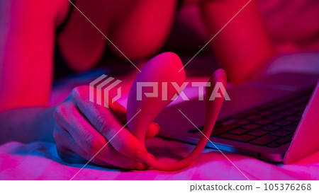 A woman is lying in bed holding a curved vibrator and looking at a laptop. Girl using sex toy in blue-red light. A woman is lying in bed holding a curved vibrator and looking at a laptop. Girl using sex toy in blue-red light. 105376268