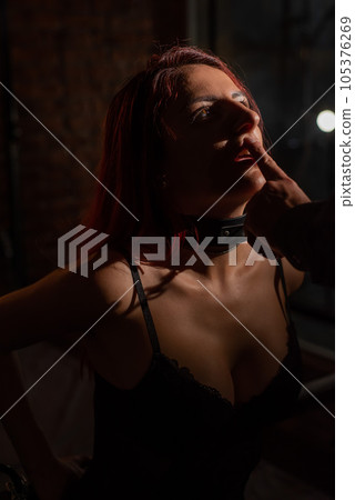 BDSM. Portrait of a red-haired woman in shackles kneeling and licking the fingers of a man. BDSM. Portrait of a red-haired woman in shackles kneeling and licking the fingers of a man. 105376269