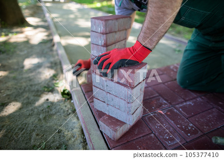 A conscientious blue-collar worker lays red paving stones in a designated area. 105377410