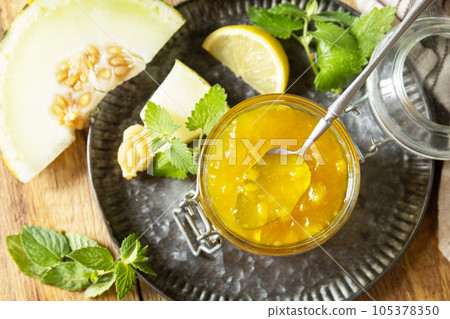 Homemade preserve. Sweet melon and citrus jam or jelly in small glass jar with fresh melon slices on wooden rustic table. View from above. 105378350