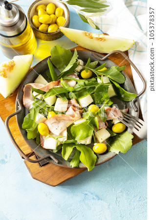 Summer mediterranean salad with melon, bacon, olives, white cheese and arugula on blue stone background. Traditional Spanish and Italian appetizer. Copy space. Summer mediterranean salad with melon, bacon, olives, white cheese and arugula on blue stone background. Traditional Spanish and Italian appetizer. Copy space. 105378357