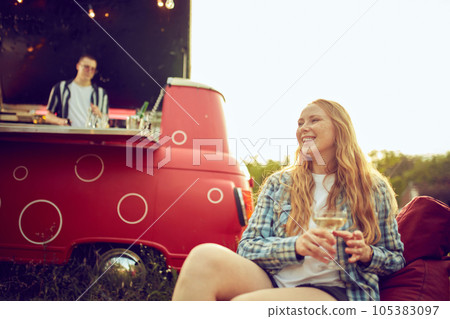 Focus on smiling, beautiful young woman sitting on floor pillow outdoors on picnic, drinking cocktails and having fun. Blurred man cooking cocktails in van 105383097