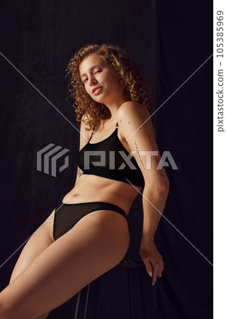 Portrait of fit woman in black underwear sitting on black chair and looking at camera on black studio background. 105385969