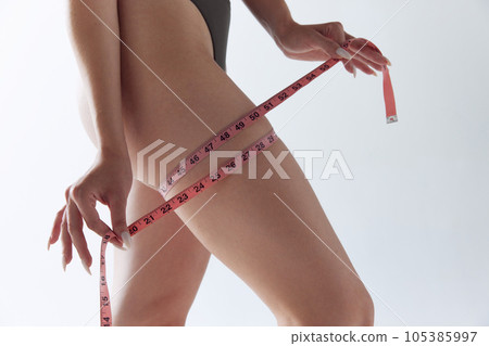Cropped image of fit young woman in underwear posing measuring leg thigh with measure tape over whitw studio background. 105385997
