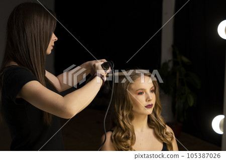 Professional stylist hairdresser curls long female hair with curling iron and makes beautiful curled strands of female hair. Professional stylist hairdresser curls long female hair with curling iron and makes beautiful curled strands of female hair. 105387026
