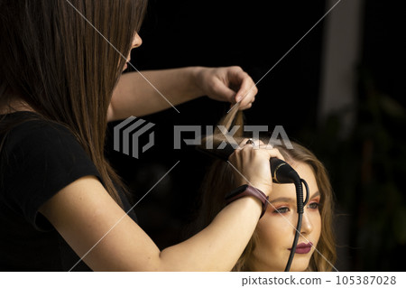 The work of the hair stylist, hairdresser with hairstyles for a beautiful young woman client. Stylist curls hair with clipless curling iron. The work of the hair stylist, hairdresser with hairstyles for a beautiful young woman client. Stylist curls hair with clipless curling iron. 105387028