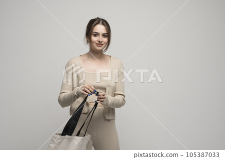 Brunette cheerful millennial woman holding white eco bag standing over white studio background. Lady holding flax shopper handbag. Fashion and ecology concept. Brunette cheerful millennial woman holding white eco bag standing over white studio background. Lady holding flax shopper handbag. Fashion and ecology concept. 105387033
