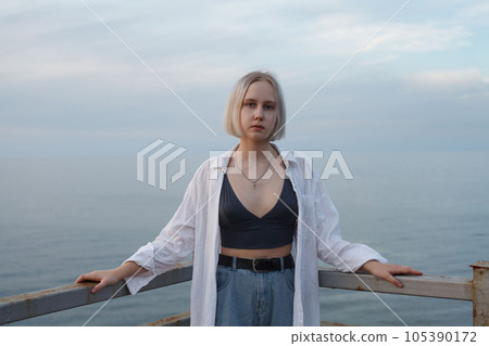 Pretty blonde woman in white shirt and tank top standing on the background of sea, cloudy sky Pretty blonde woman in white shirt and tank top standing on the background of sea, cloudy sky 105390172
